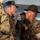 Lima Company Senior Drill Instructor Inspection The image depicts a U.S. Marine Corps drill instructor in a campaign cover (campaign hat) shouting face-to-face at a recruit during what appears to be basic training or boot camp. The drill instructor is holding a rifle and wearing a white armband, a common identifier for drill instructors. Multiple other drill instructors and recruits are visible in the background in a covered outdoor or indoor facility.