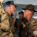 The image depicts a U.S. Marine Corps drill instructor in a campaign cover (campaign hat) shouting face-to-face at a recruit during what appears to be basic training or boot camp. The drill instructor is holding a rifle and wearing a white armband, a common identifier for drill instructors. Multiple other drill instructors and recruits are visible in the background in a covered outdoor or indoor facility.