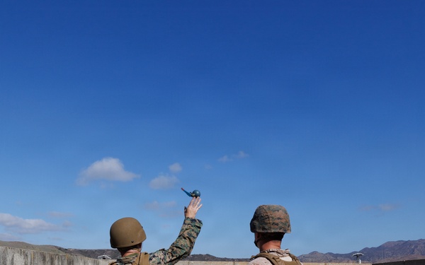 Two military personnel in combat gear are positioned against a concrete block wall, with one individual in the act of throwing what appears to be a blue training grenade over the wall. Both are wearing body armor, helmets, and camouflage uniforms consistent with U.S. Marine Corps patterns. The setting appears to be a training range in a desert environment based on the terrain and infrastructure visible in the background.