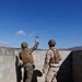 Two military personnel in combat gear are positioned against a concrete block wall, with one individual in the act of throwing what appears to be a blue training grenade over the wall. Both are wearing body armor, helmets, and camouflage uniforms consistent with U.S. Marine Corps patterns. The setting appears to be a training range in a desert environment based on the terrain and infrastructure visible in the background.