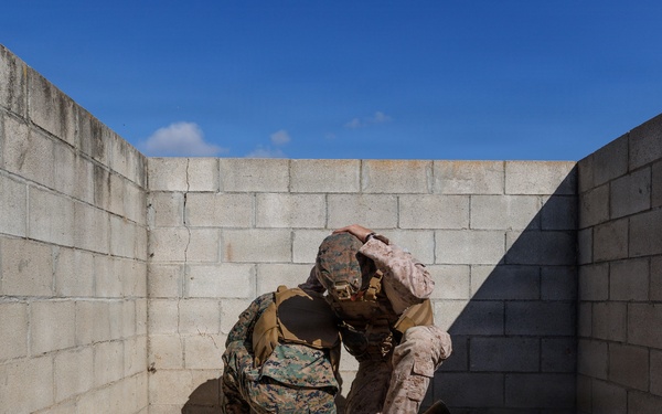 Two military personnel in camouflage uniforms are crouched together in a corner formed by concrete block walls. One individual appears to be assisting or checking on the other in what looks like a training exercise environment. The setting appears to be a military training facility with sandy ground and cinder block construction.