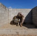 Two military personnel in camouflage uniforms are crouched together in a corner formed by concrete block walls. One individual appears to be assisting or checking on the other in what looks like a training exercise environment. The setting appears to be a military training facility with sandy ground and cinder block construction.