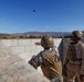 Two military personnel in combat gear are positioned behind a concrete block wall, with one individual appearing to throw or launch a small object into the air. The scene takes place at what appears to be a military training facility with desert terrain and mountains visible in the background. A small dark object is visible in mid-air above the wall, consistent with a thrown training device or grenade simulator.