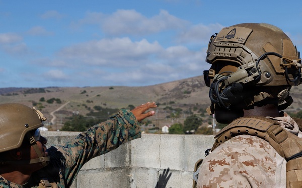 Two military personnel are positioned on what appears to be a rooftop or elevated concrete structure, with one individual pointing toward a distant location. Both are wearing tactical gear including body armor, helmets, and camouflage uniforms. The setting appears to be a training environment based on the controlled surroundings and visible terrain in the background.