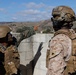Two military personnel are positioned on what appears to be a rooftop or elevated concrete structure, with one individual pointing toward a distant location. Both are wearing tactical gear including body armor, helmets, and camouflage uniforms. The setting appears to be a training environment based on the controlled surroundings and visible terrain in the background.