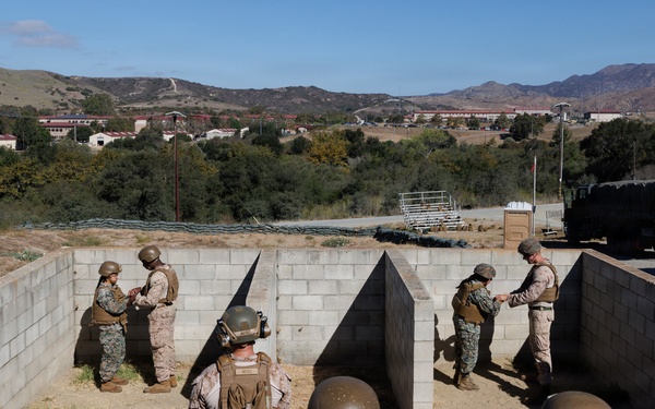 A group of U.S. Marines in combat gear and camouflage uniforms are conducting a training exercise at what appears to be a military installation, likely Camp Pendleton in California. Several Marines are positioned at concrete block wall structures that simulate urban or fortified environments, while others observe from behind. The background shows rolling hills, military vehicles, and base buildings consistent with a Southern California military training facility.