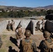 A group of U.S. Marines in combat gear and camouflage uniforms are conducting a training exercise at what appears to be a military installation, likely Camp Pendleton in California. Several Marines are positioned at concrete block wall structures that simulate urban or fortified environments, while others observe from behind. The background shows rolling hills, military vehicles, and base buildings consistent with a Southern California military training facility.