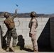 Two military personnel in combat uniforms and helmets are conducting a grenade throwing exercise at what appears to be a training facility. One soldier is in the act of throwing a blue training grenade over a concrete block wall while the other observes. The setting includes a hilly, arid landscape consistent with a military training base in the western United States.
