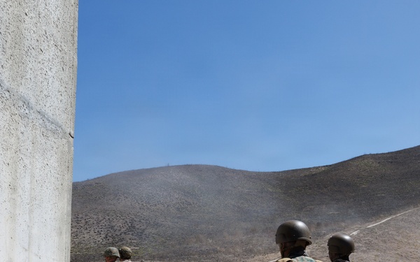 The image shows military personnel, appearing to be U.S. Marines based on their MARPAT camouflage uniforms, moving tactically toward a concrete barrier structure marked with the number '2' on an outdoor training range. The setting appears to be a dry, arid military training facility with rolling hills in the background, consistent with installations in Southern California such as Camp Pendleton. The personnel are wearing combat helmets, body armor, and tactical backpacks, suggesting a live-fire or tactical training exercise.