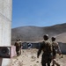 The image shows military personnel, appearing to be U.S. Marines based on their MARPAT camouflage uniforms, moving tactically toward a concrete barrier structure marked with the number '2' on an outdoor training range. The setting appears to be a dry, arid military training facility with rolling hills in the background, consistent with installations in Southern California such as Camp Pendleton. The personnel are wearing combat helmets, body armor, and tactical backpacks, suggesting a live-fire or tactical training exercise.