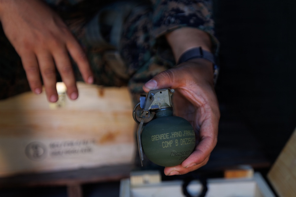 A person wearing military camouflage uniform is holding a fragmentation hand grenade. The grenade is olive drab green with visible stenciled text reading 'GRENADE, HAND, FRAG' and 'COMP B' along with a partial lot number. A wooden ammunition crate is visible in the background.