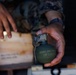 A person wearing military camouflage uniform is holding a fragmentation hand grenade. The grenade is olive drab green with visible stenciled text reading 'GRENADE, HAND, FRAG' and 'COMP B' along with a partial lot number. A wooden ammunition crate is visible in the background.