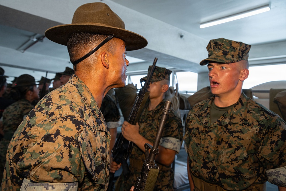 A U.S. Marine drill instructor wearing a campaign cover (drill sergeant hat) is confronting or instructing a recruit in MARPAT camouflage utilities. The scene appears to take place in an indoor training facility with multiple other Marines visible in the background. This is consistent with a military basic training or recruit depot environment.