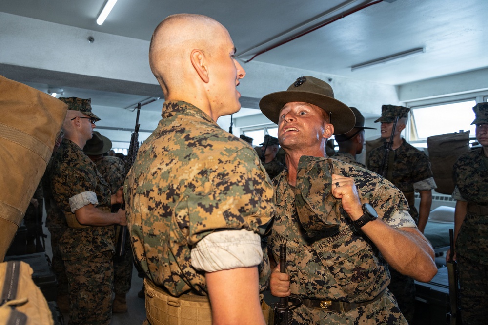 A U.S. Marine drill instructor wearing a campaign cover (drill sergeant hat) is confronting a recruit in what appears to be a barracks setting. The drill instructor is grabbing the recruit's collar and appears to be yelling, a classic training scenario during Marine Corps boot camp. Multiple other recruits and personnel are visible in the background.