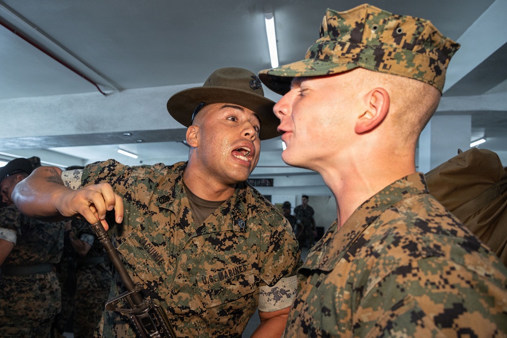 A U.S. Marine drill instructor wearing a campaign cover (drill sergeant hat) is shouting directly at a recruit in MARPAT camouflage uniform during what appears to be a training or processing event. The drill instructor is in close proximity to the recruit's face in a classic military instruction/discipline posture. The setting appears to be an indoor facility with other personnel visible in the background.