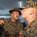 A U.S. Marine drill instructor wearing a campaign cover (drill sergeant hat) is shouting directly at a recruit in MARPAT camouflage uniform during what appears to be a training or processing event. The drill instructor is in close proximity to the recruit's face in a classic military instruction/discipline posture. The setting appears to be an indoor facility with other personnel visible in the background.