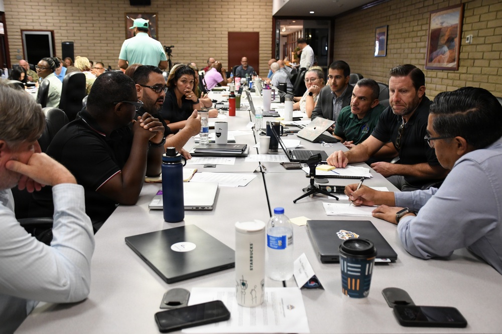 A large group of professionals are seated around a long conference table engaged in what appears to be a formal meeting or workshop. The room has a brick wall interior with framed artwork, and participants have laptops, documents, water bottles, and coffee cups spread across the table. The meeting appears to involve active discussion with multiple side conversations occurring simultaneously.