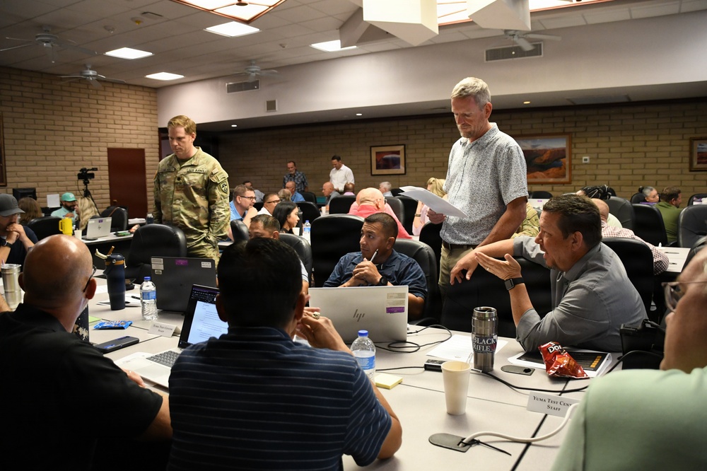 A large conference or briefing room meeting is underway, likely at a military installation based on the presence of a uniformed U.S. Army soldier and a visible nameplate reading 'Yuma Test Center Staff'. Participants, a mix of military personnel and civilians, are seated around tables with laptops, water bottles, and documents. A standing civilian man appears to be presenting or discussing information with seated colleagues.