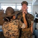 A U.S. Marine Corps drill instructor wearing a campaign cover (Smokey Bear hat) is conducting a close-up inspection or correction of a recruit in what appears to be a barracks bay. The drill instructor is pointing at or near the recruit's chin/throat area while the recruit stands at attention with mouth open. Both individuals are wearing MARPAT (Marine Pattern) digital camouflage uniforms.