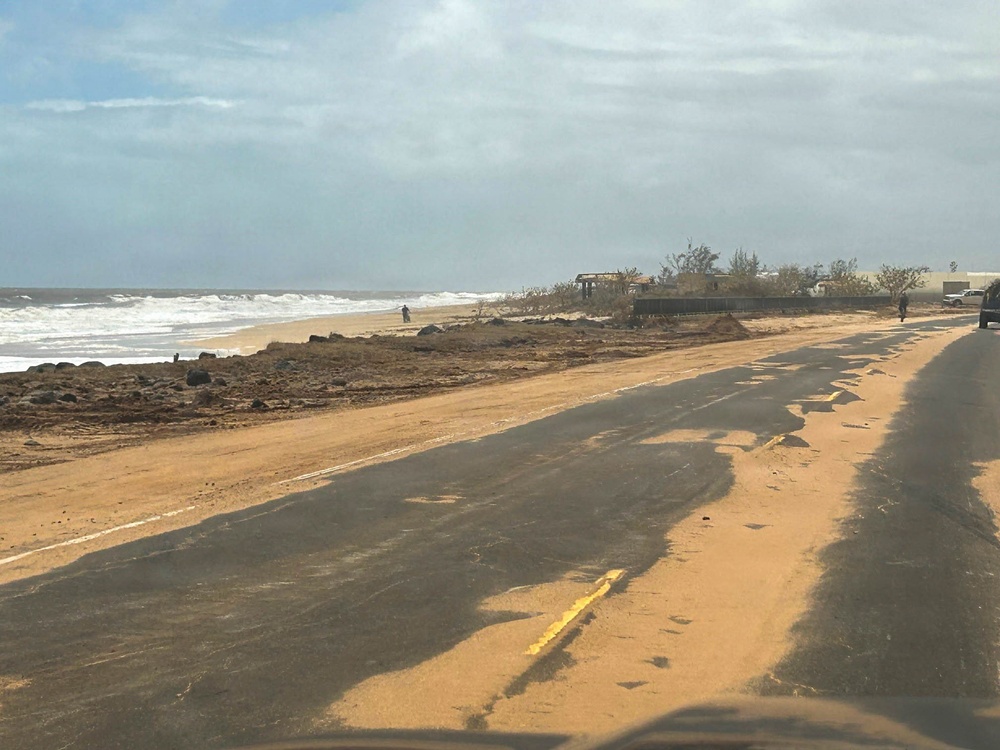 A coastal road running parallel to a beach shows significant storm or flood damage, with sand deposited across the asphalt surface and debris scattered along the shoreline. The ocean is visibly rough with active wave action, suggesting recent severe weather. Structures in the background appear damaged or partially destroyed, consistent with hurricane or tropical storm aftermath.