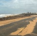 A coastal road running parallel to a beach shows significant storm or flood damage, with sand deposited across the asphalt surface and debris scattered along the shoreline. The ocean is visibly rough with active wave action, suggesting recent severe weather. Structures in the background appear damaged or partially destroyed, consistent with hurricane or tropical storm aftermath.