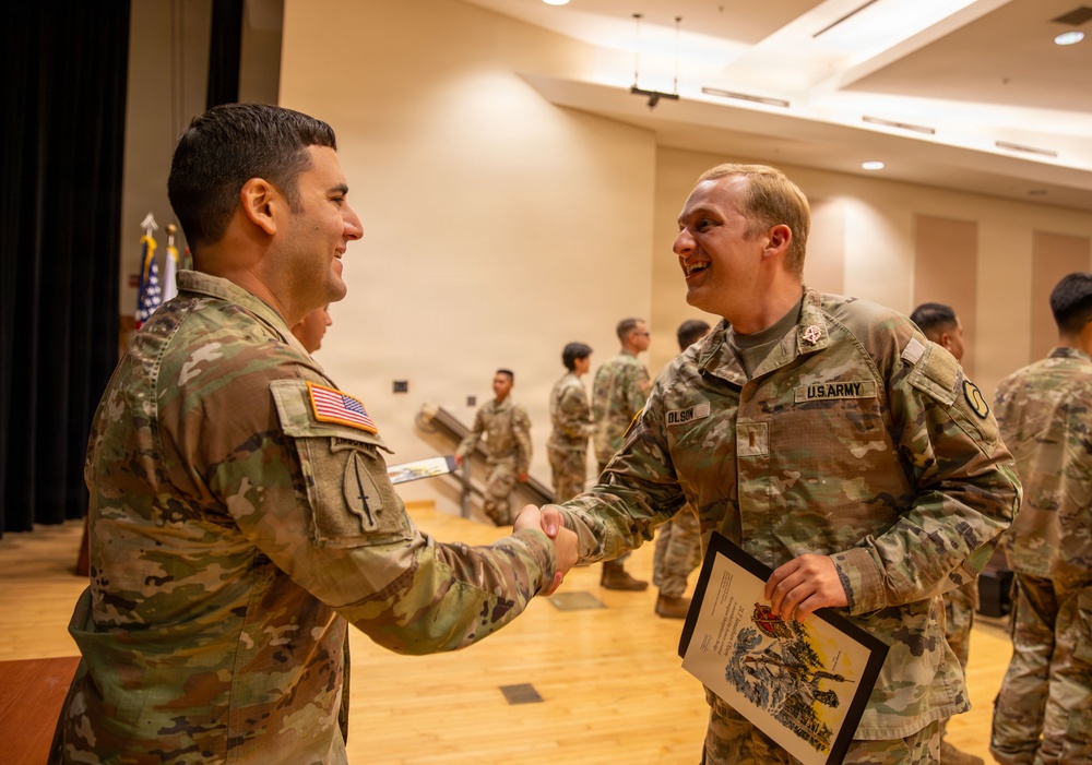 Two U.S. Army soldiers in OCP (Operational Camouflage Pattern) uniforms are shaking hands and smiling in what appears to be a formal ceremony or award presentation. The soldier on the right, identified by his name tape as 'OLSON,' is holding a framed certificate or award document. Additional soldiers are visible in the background of what appears to be a military auditorium or event hall.