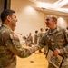 Two U.S. Army soldiers in OCP (Operational Camouflage Pattern) uniforms are shaking hands and smiling in what appears to be a formal ceremony or award presentation. The soldier on the right, identified by his name tape as 'OLSON,' is holding a framed certificate or award document. Additional soldiers are visible in the background of what appears to be a military auditorium or event hall.