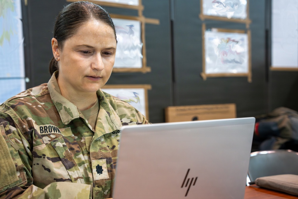 A female U.S. Army officer wearing Operational Camouflage Pattern (OCP) uniform is working on an HP laptop at a desk. Her name tape reads 'BROWN' and she displays a Lieutenant Colonel oak leaf cluster rank insignia on her chest. She appears focused on her work in what looks like a military operations or briefing room.