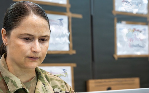 A female U.S. Army officer wearing Operational Camouflage Pattern (OCP) uniform is working on an HP laptop at a desk. Her name tape reads 'BROWN' and she displays a Lieutenant Colonel oak leaf cluster rank insignia on her chest. She appears focused on her work in what looks like a military operations or briefing room.