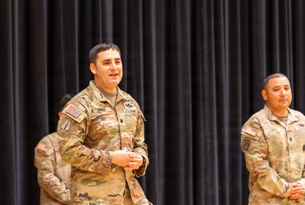 Three U.S. Army soldiers in OCP (Operational Camouflage Pattern) uniforms are standing in front of a black curtain backdrop, likely on a stage during a military ceremony or event. The central figure, identified by name tape as 'ZIMMER,' appears to be speaking or addressing an audience. A second soldier on the right is identified as 'LOPEZ.'
