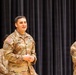 Three U.S. Army soldiers in OCP (Operational Camouflage Pattern) uniforms are standing in front of a black curtain backdrop, likely on a stage during a military ceremony or event. The central figure, identified by name tape as 'ZIMMER,' appears to be speaking or addressing an audience. A second soldier on the right is identified as 'LOPEZ.'