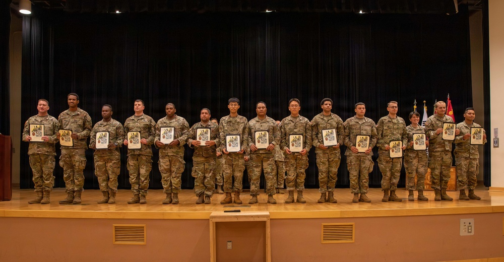 A group of approximately 14 military personnel in U.S. Army OCP (Operational Camouflage Pattern) uniforms are standing in a line on a stage, each holding what appears to be a certificate or award plaque. The setting appears to be a formal military recognition or awards ceremony held in an auditorium or multipurpose hall. Military flags are visible in the background to the right side of the stage.
