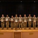 A group of approximately 14 military personnel in U.S. Army OCP (Operational Camouflage Pattern) uniforms are standing in a line on a stage, each holding what appears to be a certificate or award plaque. The setting appears to be a formal military recognition or awards ceremony held in an auditorium or multipurpose hall. Military flags are visible in the background to the right side of the stage.