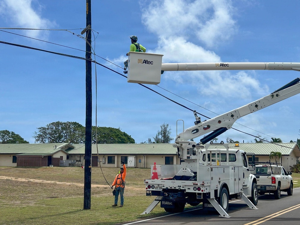 Utility workers are performing maintenance or repair work on overhead power lines using an Altec brand bucket truck. One worker is elevated in the aerial lift bucket near the utility pole and lines, while a second worker in an orange safety vest stands on the ground below. The scene takes place on a road adjacent to what appears to be a residential or institutional complex with green metal roofs.