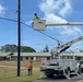 Utility workers are performing maintenance or repair work on overhead power lines using an Altec brand bucket truck. One worker is elevated in the aerial lift bucket near the utility pole and lines, while a second worker in an orange safety vest stands on the ground below. The scene takes place on a road adjacent to what appears to be a residential or institutional complex with green metal roofs.