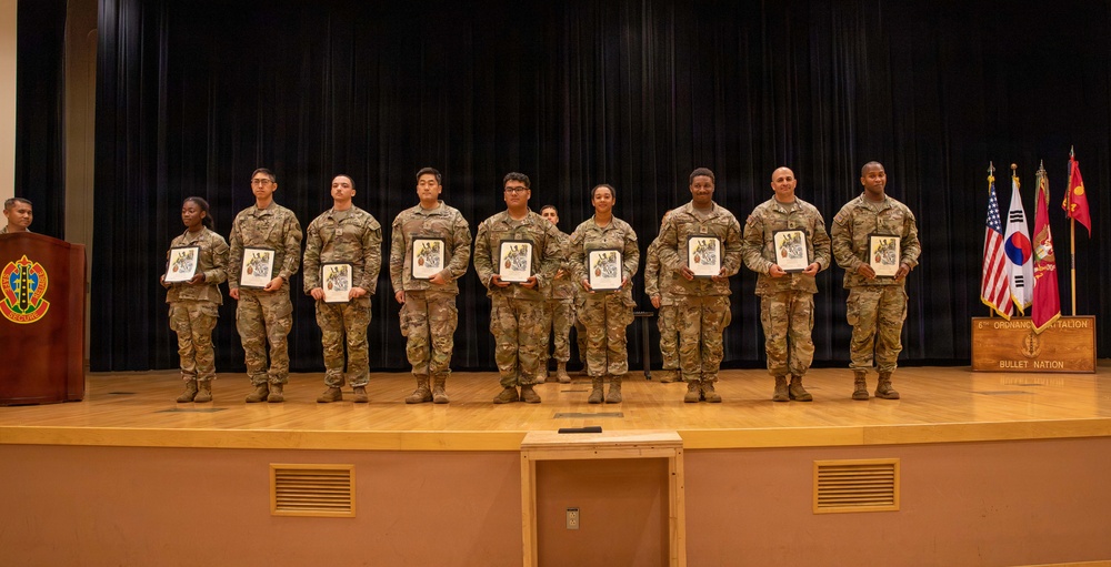A military awards or recognition ceremony taking place on a stage, with nine soldiers in Army Combat Uniforms (ACU) standing in a line holding framed certificates or plaques. The event appears to be associated with the 4th Ordnance Battalion, known as 'Bullet Nation,' as indicated by the sign on the stage. Three flags are visible in the background, including the US flag, South Korean flag, and a unit flag, suggesting this is taking place in South Korea.