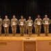 A military awards or recognition ceremony taking place on a stage, with nine soldiers in Army Combat Uniforms (ACU) standing in a line holding framed certificates or plaques. The event appears to be associated with the 4th Ordnance Battalion, known as 'Bullet Nation,' as indicated by the sign on the stage. Three flags are visible in the background, including the US flag, South Korean flag, and a unit flag, suggesting this is taking place in South Korea.