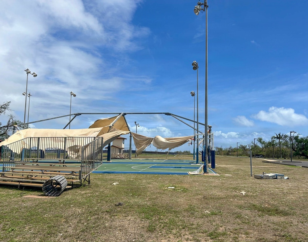 An outdoor basketball court has sustained significant storm damage, with a shade canopy structure torn and collapsed over the playing surface. Debris is scattered across the surrounding grass area, and bleachers appear displaced or damaged. The scene is consistent with post-hurricane or severe wind event destruction.
