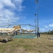 An outdoor basketball court has sustained significant storm damage, with a shade canopy structure torn and collapsed over the playing surface. Debris is scattered across the surrounding grass area, and bleachers appear displaced or damaged. The scene is consistent with post-hurricane or severe wind event destruction.