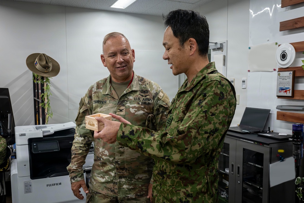 Two military personnel in camouflage uniforms are engaged in what appears to be a gift exchange or presentation ceremony in an office setting. The individual on the left is identified by name tape as 'CARTER' and wears a U.S. Army uniform, while the individual on the right wears a Japanese Ground Self-Defense Force (JGSDF) uniform. Both are examining a small wooden box, suggesting a bilateral military exchange or courtesy visit.