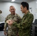 Two military personnel in camouflage uniforms are engaged in what appears to be a gift exchange or presentation ceremony in an office setting. The individual on the left is identified by name tape as 'CARTER' and wears a U.S. Army uniform, while the individual on the right wears a Japanese Ground Self-Defense Force (JGSDF) uniform. Both are examining a small wooden box, suggesting a bilateral military exchange or courtesy visit.