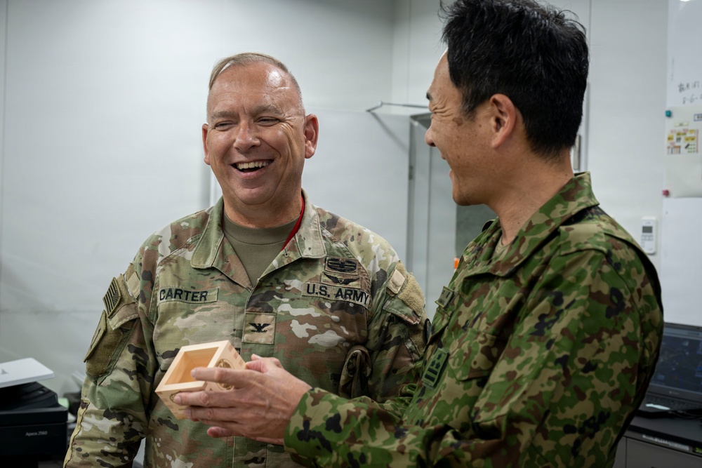 A U.S. Army Colonel named Carter is laughing and interacting with a military officer wearing what appears to be Japan Ground Self-Defense Force camouflage uniform. The two are engaged in what appears to be a gift exchange or presentation, with Carter holding a small wooden box. The atmosphere is friendly and informal, suggesting a bilateral military engagement or courtesy visit.