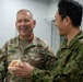 A U.S. Army Colonel named Carter is laughing and interacting with a military officer wearing what appears to be Japan Ground Self-Defense Force camouflage uniform. The two are engaged in what appears to be a gift exchange or presentation, with Carter holding a small wooden box. The atmosphere is friendly and informal, suggesting a bilateral military engagement or courtesy visit.