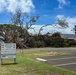 A large tree has been uprooted and fallen, likely due to a severe storm or high winds, in the parking lot area of a Navy MWR Community Recreation Center. The fallen tree's root system is fully exposed and the canopy sprawls across the ground near the facility's signage. The building in the background, which features solar panels on its roof, appears undamaged.