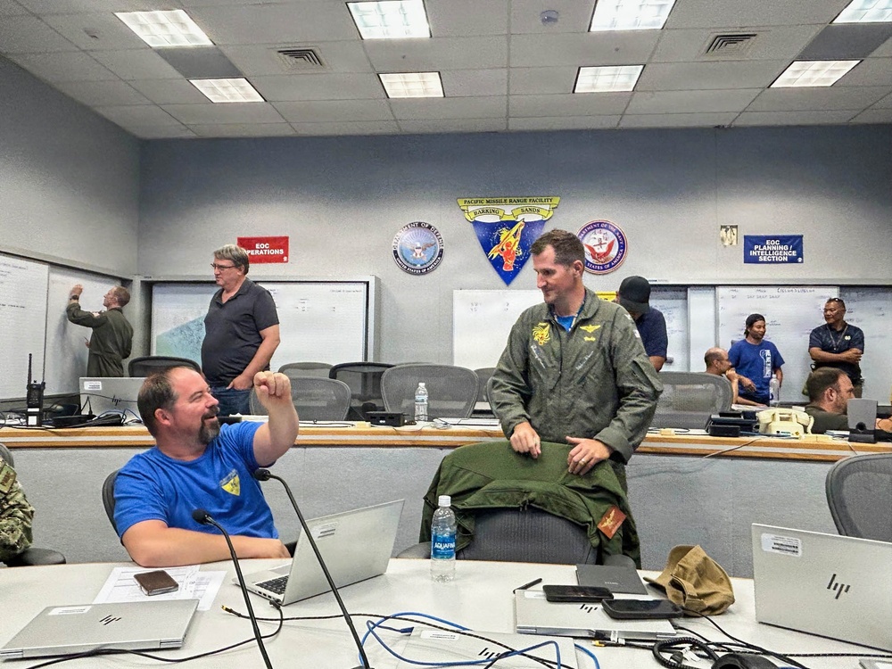 The image shows a military Emergency Operations Center (EOC) at the Pacific Missile Range Facility (PMRF) Barking Sands, with multiple personnel engaged in planning and coordination activities. A man in a blue shirt is seated at a workstation conversing with a person in a green military flight suit, while others work at whiteboards and computer stations in the background. The room displays official military insignia and is organized into operational sections.