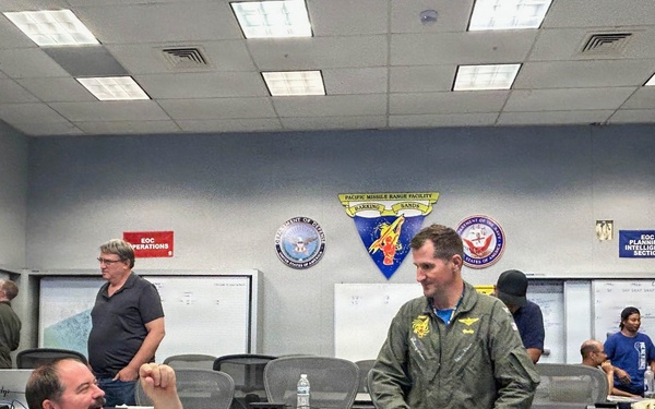 The image shows a military Emergency Operations Center (EOC) at the Pacific Missile Range Facility (PMRF) Barking Sands, with multiple personnel engaged in planning and coordination activities. A man in a blue shirt is seated at a workstation conversing with a person in a green military flight suit, while others work at whiteboards and computer stations in the background. The room displays official military insignia and is organized into operational sections.