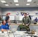 The image shows a military Emergency Operations Center (EOC) at the Pacific Missile Range Facility (PMRF) Barking Sands, with multiple personnel engaged in planning and coordination activities. A man in a blue shirt is seated at a workstation conversing with a person in a green military flight suit, while others work at whiteboards and computer stations in the background. The room displays official military insignia and is organized into operational sections.