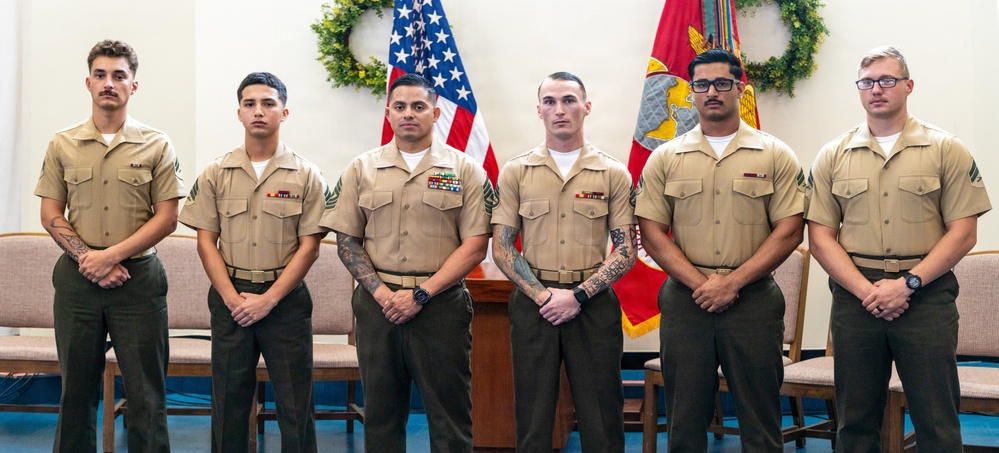 Six U.S. Marines in service uniforms (tan/khaki shirts with olive green trousers) are posed in a formal group photo. They are standing in front of an American flag and a U.S. Marine Corps flag, suggesting an official ceremony or recognition event. The individuals display varying ranks indicated by their collar insignia and ribbon/medal arrangements.