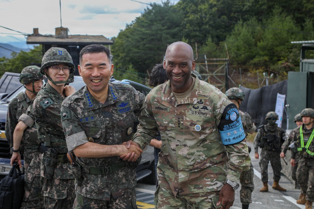 Two senior military officers — one in Republic of Korea Army (ROKA) uniform and one in U.S. Army OCP uniform — are shaking hands and smiling in what appears to be a military installation or field environment. The U.S. officer's name tape reads 'BRUNSON' and he wears a blue UN Command armband. Multiple soldiers in combat gear are visible in the background.