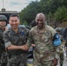 Two senior military officers — one in Republic of Korea Army (ROKA) uniform and one in U.S. Army OCP uniform — are shaking hands and smiling in what appears to be a military installation or field environment. The U.S. officer's name tape reads 'BRUNSON' and he wears a blue UN Command armband. Multiple soldiers in combat gear are visible in the background.